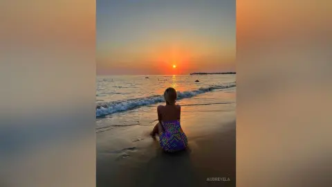 Woman in a colorful patterned sundress sitting on a beach at sunset watching the ocean waves.