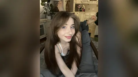 Smiling young woman with long brown hair in a white tank top relaxing on a sofa in a cozy living room.