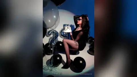 Glamorous woman in a black dress holding a small cake amidst silver balloons in a moody studio setting.