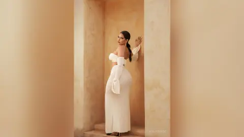 Elegant woman in a white off-the-shoulder gown posing in a minimalist beige architectural setting.