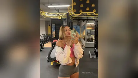 Young woman in pink sports bra and white jacket taking a selfie in a modern gym setting.