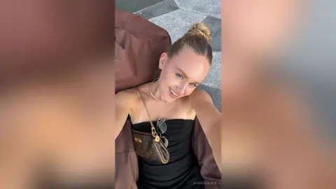 BonnyWalace smiles for a selfie wearing a black strapless top and a gold necklace while relaxing outdoors.