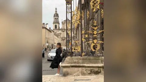 Stylish woman in a black coat walking past ornate gold gates on a historic European city street.
