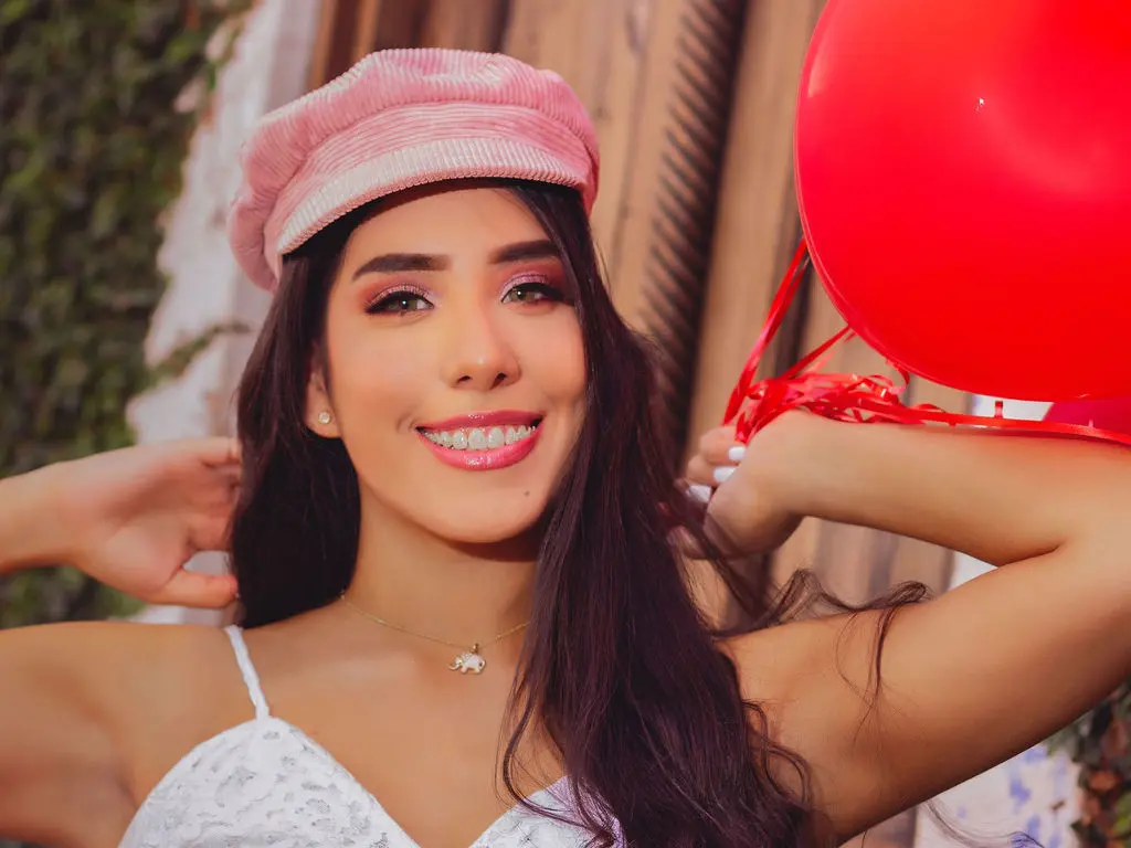 Smiling woman in a white lace top and pink beret holding a red balloon in a sunny outdoor setting.