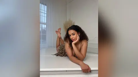 Young woman in a leopard print dress relaxing on a white surface in a bright, minimalist room.
