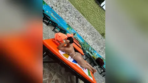 Fit woman in white bikini relaxing on an orange lounge chair at a sunny outdoor tropical resort garden.