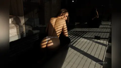 DelanaGrace poses naked in a black bathtub, showing her bare back and buttocks under slatted shadows.