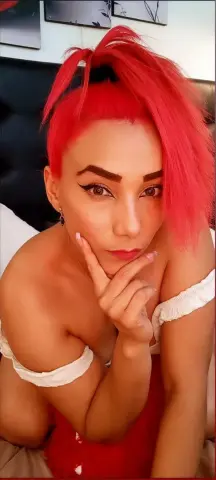 Woman with bright red hair in a white off-the-shoulder top posing thoughtfully in a bedroom setting.