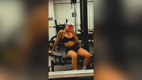 Athletic woman in black sports bra and shorts sitting on gym equipment for a fitness selfie.