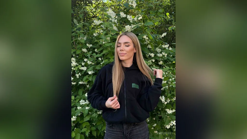 AmiliRay — AmiliRay stands in front of green bushes and white flowers wearing a black hoodie and dark jeans.
