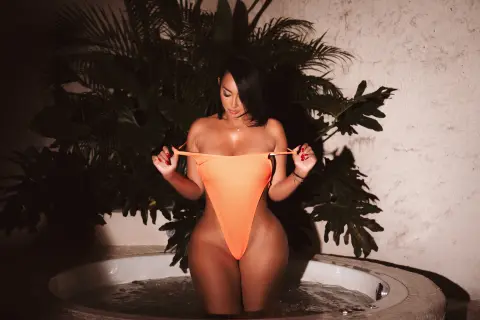 Woman in orange one-piece swimsuit sitting in a luxury hot tub against tropical palm leaves in moody lighting.