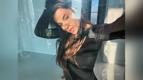AnaMathers poses against a glass window, wearing a black long-sleeved top with her long brown hair falling over her shoulder.