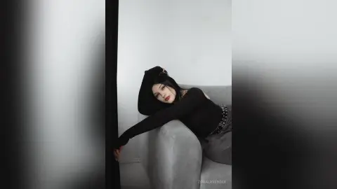 Moody portrait of a woman in a black long-sleeve top relaxing on a grey sofa in a minimalist room.