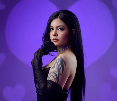 Moody portrait of a woman with long black hair and tattoos wearing black gloves against a purple heart background.