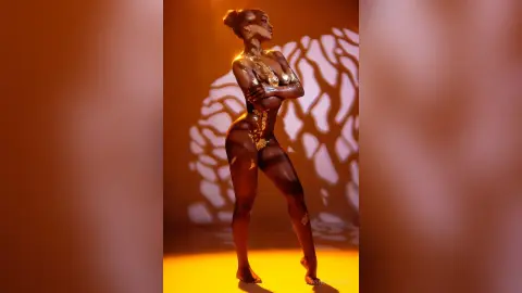 Elegant Black woman in gold body paint posing against warm shadows in a moody, golden studio setting.