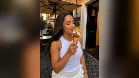 FabianaLima holds a chocolate ice cream cone in a white tank top while standing outdoors near a cafe table and umbrella.