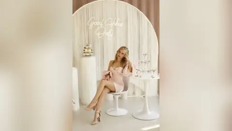 StellaFletcher poses in a light pink dress while sitting on a white chair next to a small table in a bright studio setting.