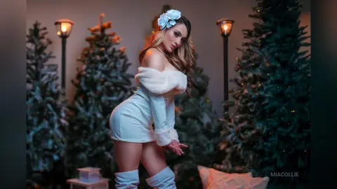 Glamorous woman in a white fuzzy mini dress and flower headband posing in a festive Christmas tree setting.