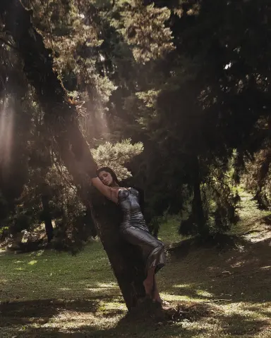 Elegant woman in a silver satin dress leaning against a tree in a sunlit, moody forest setting.