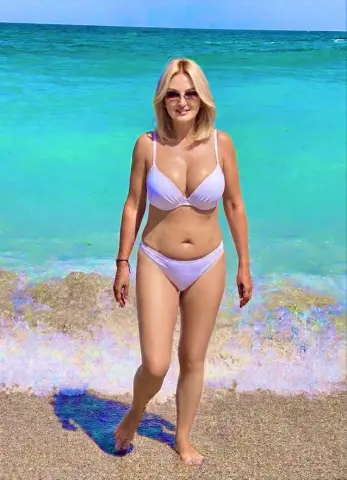 Blonde woman in a white bikini standing on a sunny tropical beach with turquoise ocean waves.