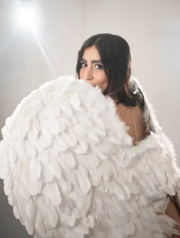 Beautiful woman in a white feathered angel costume posing in a dreamy, bright studio setting.