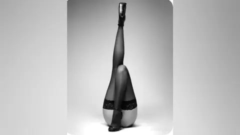 Elegant woman in black sheer tights and high heels posing in a minimalist black and white studio setting.