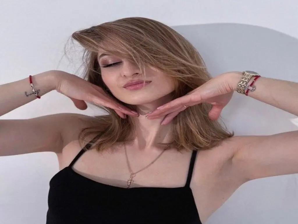 Blonde woman in a black tank top posing against a white wall with a relaxed, serene expression.