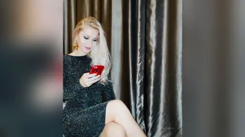 Blonde woman in a sparkling black cocktail dress using a smartphone against gold curtains.