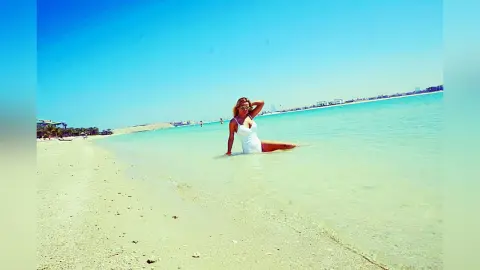 MilenaBia wades in clear turquoise ocean water wearing a white one-piece swimsuit on a sunny beach.