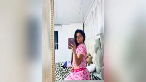 Young woman in pink heart print pajamas taking a mirror selfie in a bright bedroom.