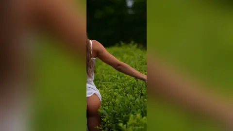Slim woman in a white summer dress standing by lush green garden bushes in soft natural light.