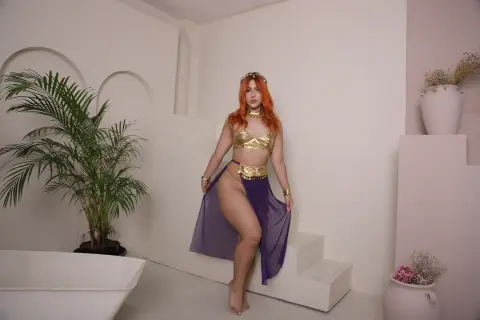 Woman in gold crop top and purple flowing skirt posing in a bright minimalist room with tropical plants.