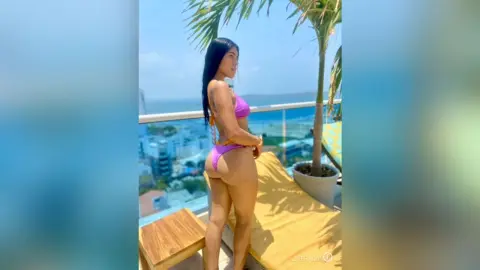 Young woman in a purple bikini relaxing on a sunny rooftop balcony overlooking a coastal city view.