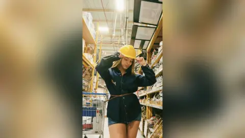 Young woman in a navy denim shirt and yellow cap smiling in a bright warehouse setting.