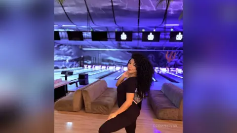 Smiling woman in a black outfit posing in a modern neon-lit bowling alley lounge with blue mood lighting.