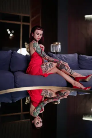 Tattooed woman in a red dress sitting on a blue sofa in a moody, dimly lit modern living room.