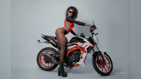 Stylish woman in red bodysuit and sheer tights posing with a black and orange sport motorcycle in a studio.