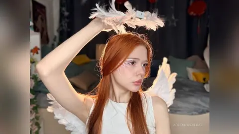 Redheaded woman in white angel costume with feathered wings and headband in a cozy bedroom setting.
