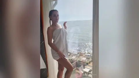 Young woman in a white towel standing by a balcony window overlooking the sunny ocean view.