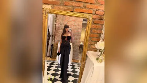 LanaRhode poses in a mirror wearing a long black cutout dress, standing on a checkered floor in front of a brick wall.