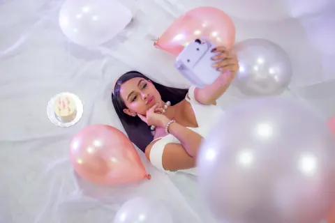 Young woman in white dress lying amidst pink and white balloons with a birthday cake in a dreamy celebratory mood.