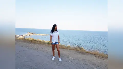 Young woman in white t-shirt and denim shorts standing on a coastal cliff overlooking the blue ocean.