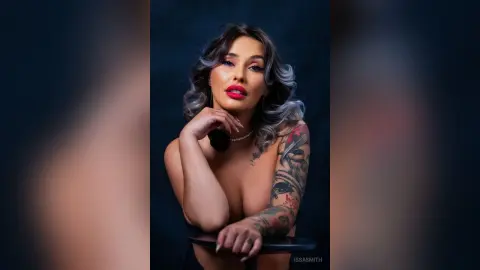 Glamorous woman with wavy hair and tattoos wearing a pearl necklace in a moody studio portrait.