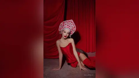 Elegant woman in a red corset and ornate white headdress posing against a dramatic red curtain backdrop.