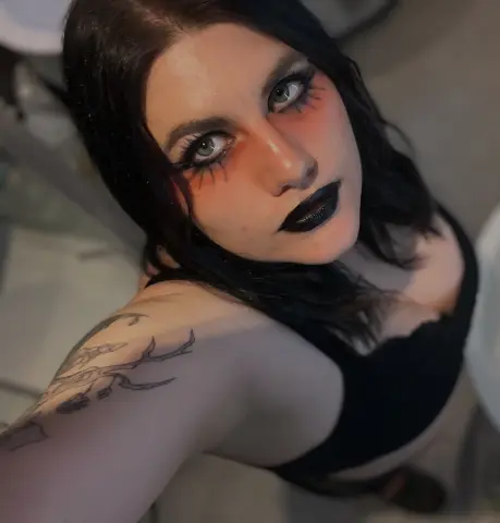 Woman with dark hair and gothic makeup wearing a black tank top in a moody indoor setting.