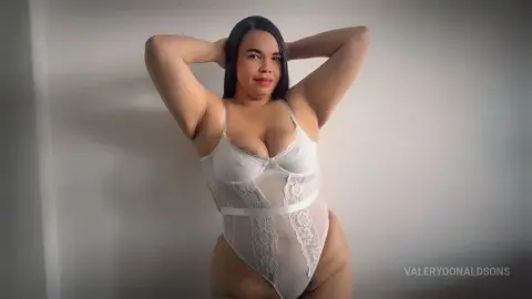 ValeryDonaldsons poses in a white lace bodysuit with her hands behind her head against a plain wall.