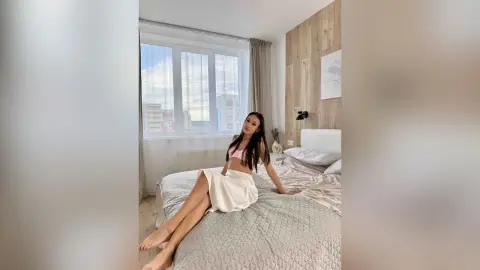ChloeTeles sits on a bed in a bright room, wearing a pink bra and a white skirt.