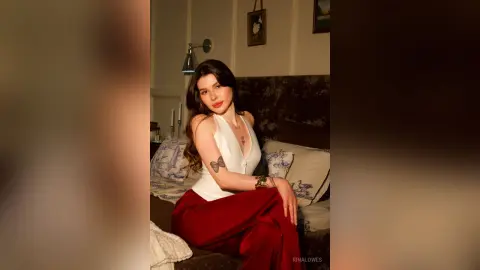 Young woman in a white sleeveless top and red trousers sitting on a bed in a cozy, dimly lit bedroom.