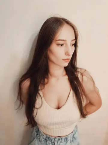 Young woman with long brown hair wearing a white ribbed tank top against a neutral wall in soft lighting.