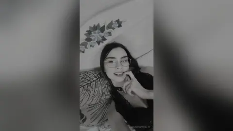Smiling young woman with glasses relaxing in bed for a cozy, moody black and white portrait.